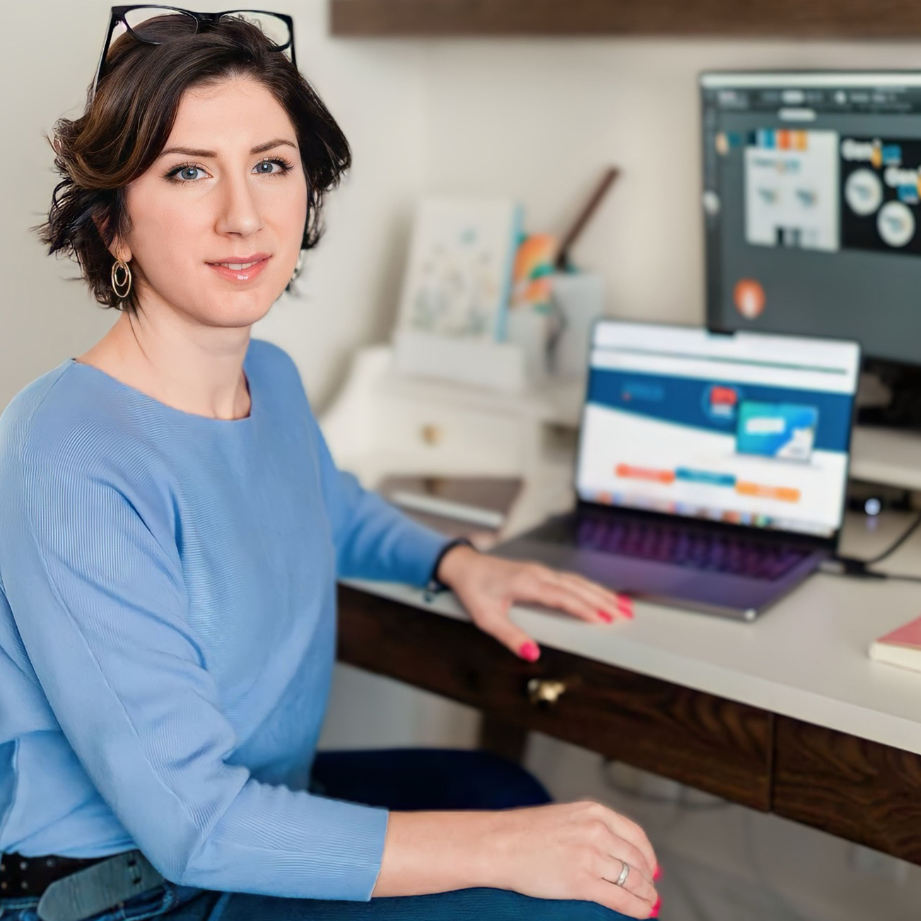 Woman sitting at desk with computer in the background. She is wearing glasses on top of her head, has short brown hair and is wearing a blue shirt. 